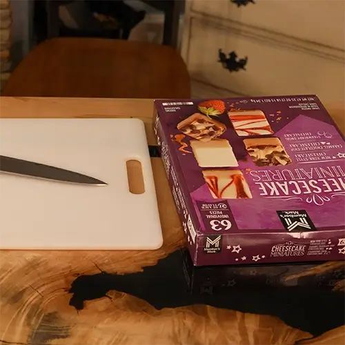 A box of cheesecake bites sitting next to a cutting board with a knife.