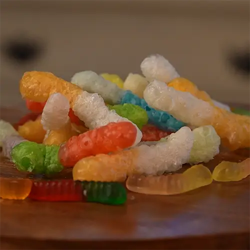 A stack of large freeze dried gummy worms puffed up and airy surrounded by several original gummy worms to show the difference in size and texture.