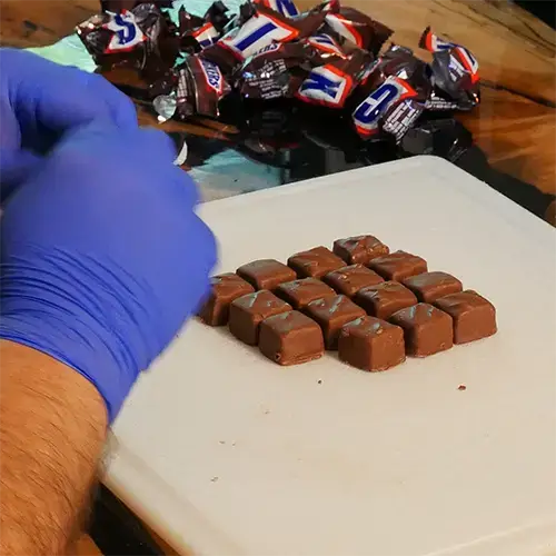 A man wearing blue nitrile gloves opening up 16 Snicker minis and placing them neatly on a cutting board.