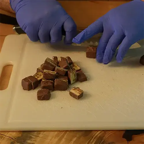 A man wearing blue nitrile gloves cutting Snickers minis in half using a kitchen knife and cutting board.