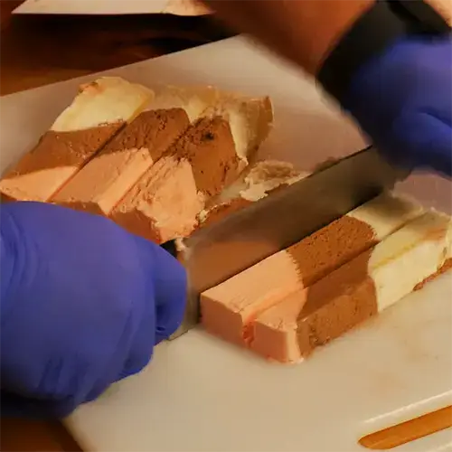 A person with blue nitrile gloves cutting frozen slices of Neapolitan ice cream into bars.