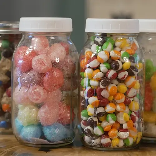 Several mason jars filled with freeze dried candies, from left to right: caramel M&M's, Jolly Ranchers, Skittles, and Gummy Worms.