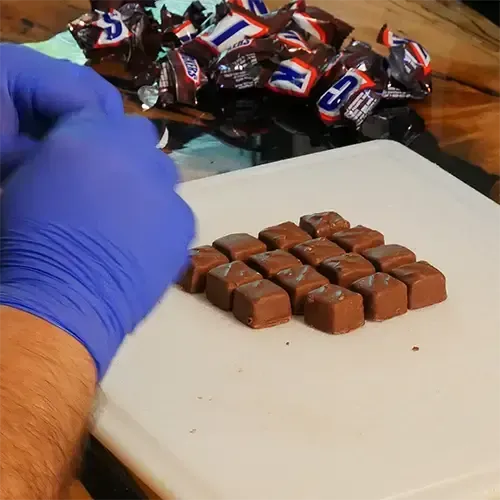 A man wearing blue nitrile gloves opening up 16 Snicker minis and placing them neatly on a cutting board.