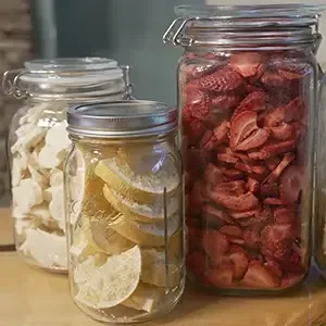 A selection of glass jars containing freeze-dried strawberries, orange segments, and banana pieces, arranged on a wooden table.